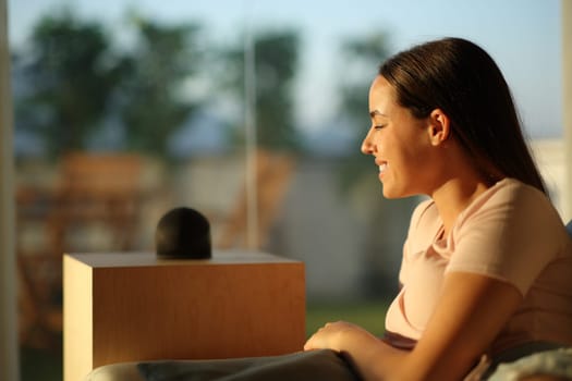 Happy woman with smart speaker sitting on a couch at sunset at home