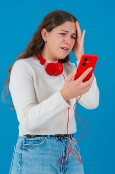 Caucasian woman looks shocked while reading bad news on her smartphone. She wears headphones and a white sweater, reacting with concern against a solid blue background.