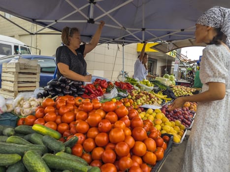Vegetable Street market. Woman selling fresh vegetables and fruits to a customer under a blue tent on a bright day in Sokuluk, Kyrgyzstan - June 28, 2024