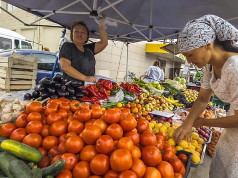 Vegetable Street market. Woman selling fresh vegetables and fruits to a customer under a blue tent on a bright day in Sokuluk, Kyrgyzstan - June 28, 2024