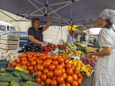 Vegetable Street market. Woman selling fresh vegetables and fruits to a customer under a blue tent on a bright day in Sokuluk, Kyrgyzstan - June 28, 2024