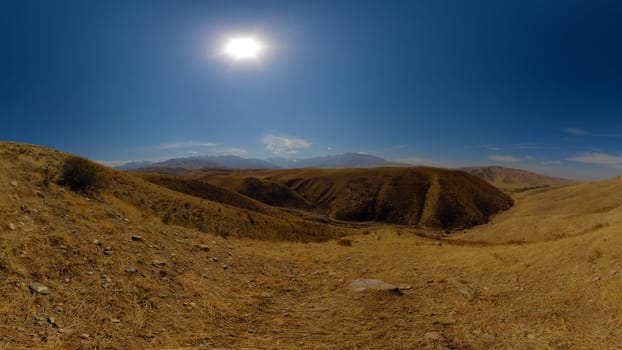 A wide panoramic view of arid, yellow-brown rolling hills under a clear blue sky with the sun shining.