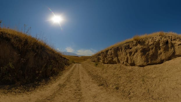 A dirt road or track cuts through dry, yellow-brown hills under a clear blue sky with the sun shining.