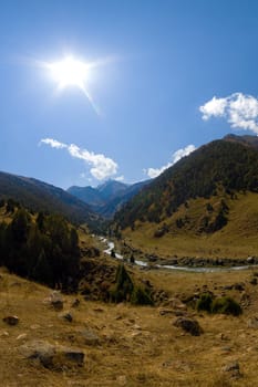 Elevated panoramic view of a sunlit mountain valley. A river winds between grassy foreground slopes and distant forested mountains under a blue sky in Zhylamysh gorge, Kyrgyzstan