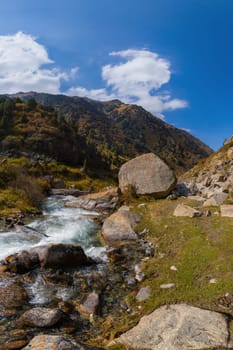 A clear mountain stream with small rapids flows through a valley filled with rocks and patches of grass and low shrubs.