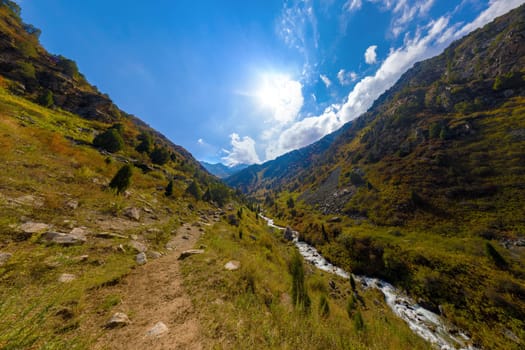 Panoramic high angle view down steep Zhylamysh Gorge. A river rushes below slopes covered with grass and juniper trees under a bright, cloudy sky.