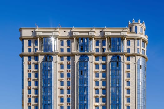 A neoclassical high-rise in Kyrgyzstan featuring a beige stone facade, arched windows, vertical columns, and reflective blue glass panels under a clear sky, blending traditional and modern architectural elements.