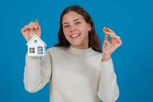 Happy young woman holding a house model and keys, expressing excitement. She wears a white sweater and stands against a blue background. Concept of home ownership, real estate, or investment.