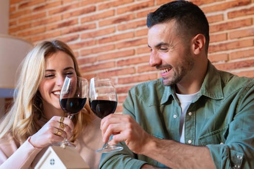 Smiling couple toasting with red wine while celebrating their new home. A miniature house and keys on the table symbolize the beginning of a new chapter in their life together.