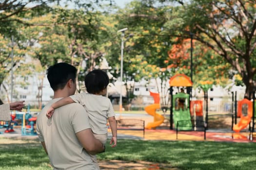 Father carries his toddler while walking toward a vibrant play area in a park.