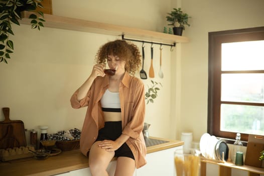 Relaxed young woman enjoys a hot beverage sitting on a kitchen counter.