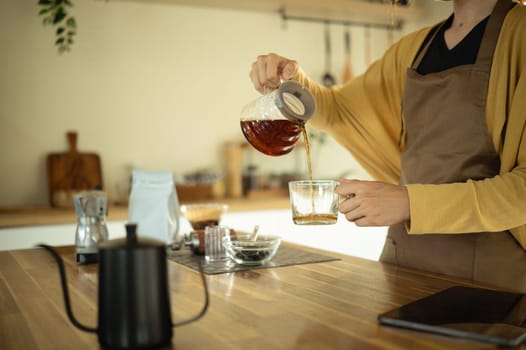 Close up of a woman in an apron pouring hot brewed coffee into a transparent glass mug.