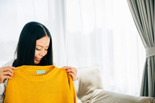 Excited Asian woman unpacks a shirt from a cardboard box smiling at home. Delighted customer unboxes online shopping package showcasing new clothes. Delivery and shopping concept.