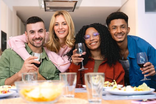 Group of multiethnic friends smiling at the camera while enjoying wine and dinner together at home. A joyful moment of connection, laughter, and celebration around a cozy and inviting table.