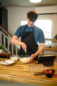 Smiling young man in apron whisking pancake batter in kitchen.