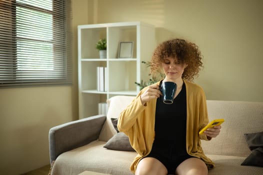 Young woman sitting on a cozy couch, sipping from a mug and using smartphone in a stylish home interior.