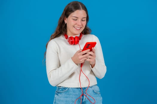 Happy young woman wearing red headphones around her neck while using her red smartphone. She smiles as she types a message, standing against a bright blue background in a casual moment.