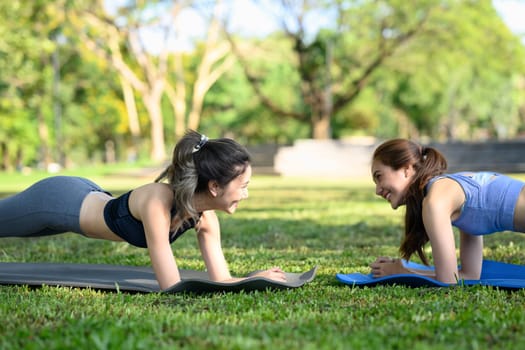 Happy female friends doing plank exercises on yoga mats in the park.