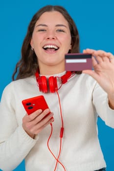 Happy young woman in a white sweater and red headphones smiling while showing a credit card to the camera and holding a red smartphone, against a blue background, symbolizing online shopping. Vertical