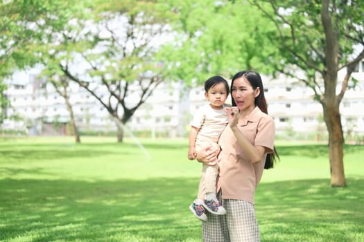 Asian mother and her toddler stroll through a lush green park.