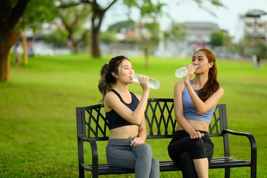 Sporty women sit on a bench and drink water, enjoying a refreshing break after exercise.