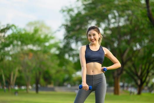 Portrait of a cheerful female athlete lifting dumbbells in a green park.