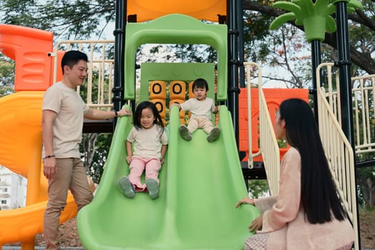Smiling siblings enjoy a fun ride down a double slide as their parents stand by.