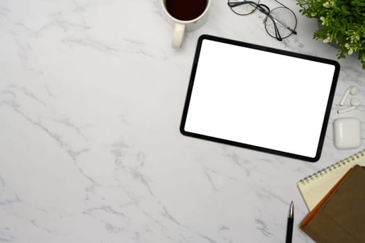 Flat lay of digital tablet with blank screen, coffee mug, notebook, and glasses on marble desk.