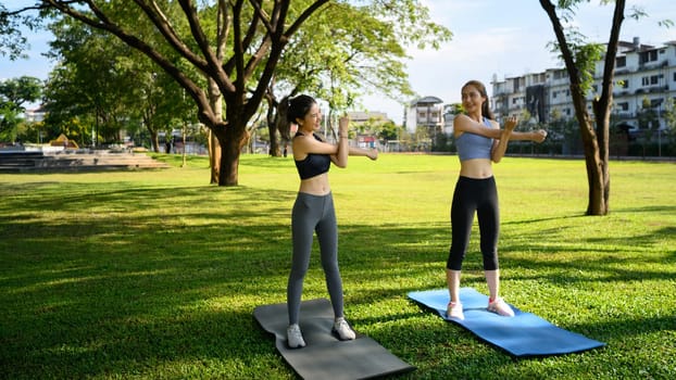 Two athletic women stretch their arms on yoga mats in a sunny park.
