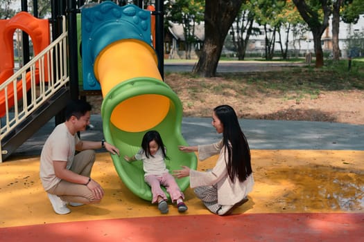 Joyful little girl enjoys sliding with support from her loving parents on both sides.