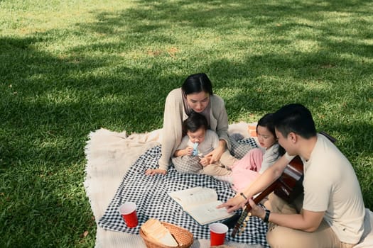 Parents read a book to their young children while relaxing on a blanket outdoors.