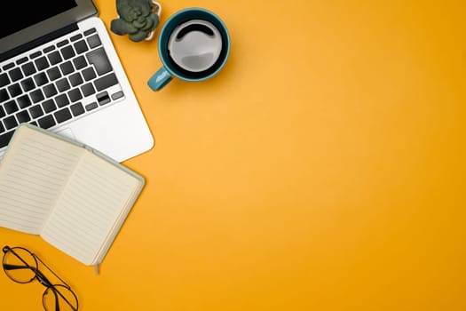 Creative workspace with laptop, notebook, eyeglasses, and coffee mug on yellow background.