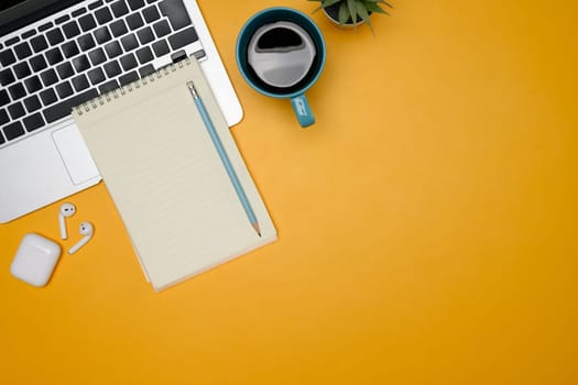 Creative workspace with laptop, notebook, coffee mug and wireless earbuds on yellow background.
