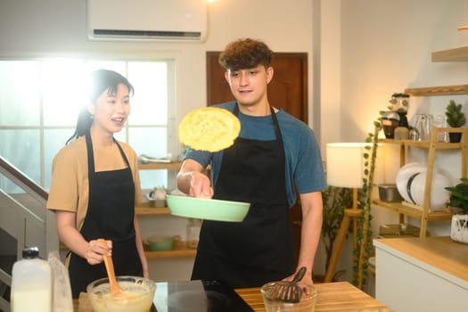 Playful couple flipping pancake, sharing laughter during home cooking.