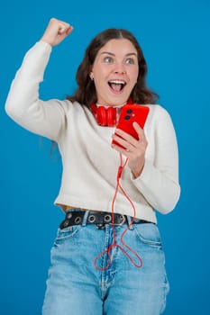 Happy young woman wearing red headphones around her neck, clenching her fist in excitement while looking at her smartphone. Her joyful reaction stands out against the blue background.Vertical