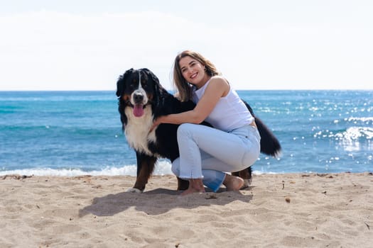 Portrait of a cheerful young woman kneeling beside her dog on a sandy beach. Both look at the camera on a sunny day, enjoying a peaceful moment by the sea with joy, friendship, and nature.