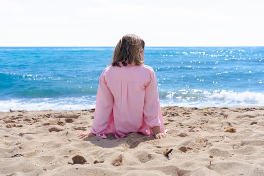 Rear view of a young woman sitting alone on the sandy beach, looking out at the calm ocean. Dressed in a pink shirt, she enjoys a quiet and relaxing moment in nature under the bright sun.