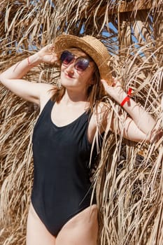 Woman on vacation near straw umbrella on beach. summer holiday concept. woman near palm leaves. person in a one piece swimsuit and sun hat
