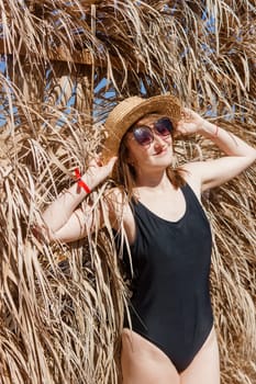 Woman on vacation near straw umbrella on beach. summer holiday concept. woman near palm leaves. person in a one piece swimsuit and sun hat