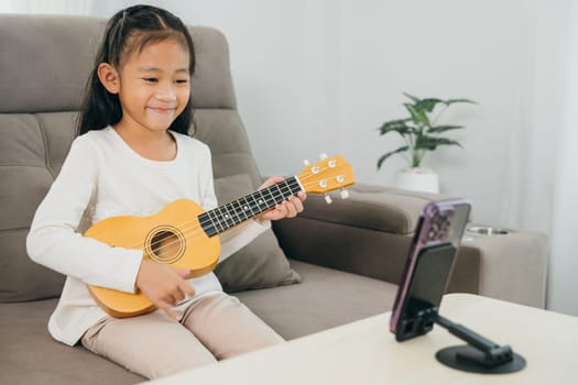 A young girl sits on a couch playing a ukulele, with a smartphone on a stand streaming her online music lesson. The scene highlights creative learning at home with modern digital tools.