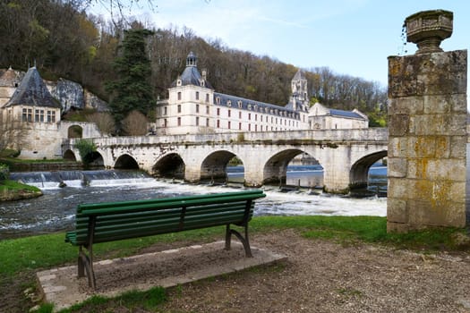 A green bench sits on a lawn near a bridge. It is located near a body of water, crossed by the bridge. Brandonne, France