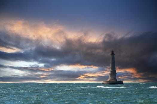 A lighthouse is seen in the distance on a cloudy day. The sky is filled with clouds, and the water is choppy. Scene is somewhat ominous, as the lighthouse stands tall against the stormy sky