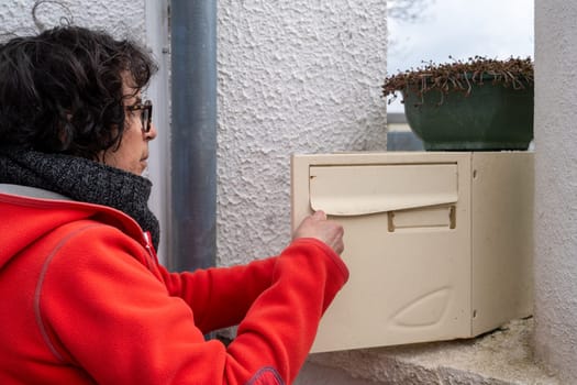 A woman in a red coat opens a mailbox. The box is hanging on the wall.