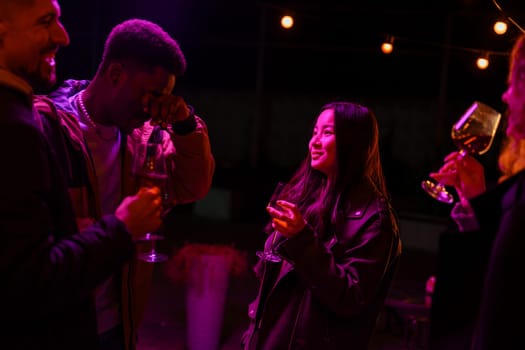 Four friends share laughter and drinks at a lively outdoor party, illuminated by colorful string lights, creating a warm atmosphere during the night.