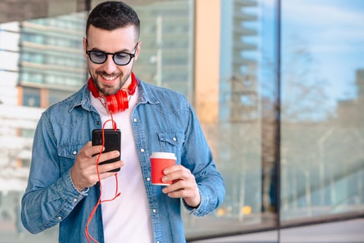 Young man with red headphones enjoying coffee while using his smartphone in a city environment. He smiles while reading or texting, standing outside modern glass buildings in a casual moment.
