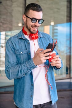 Young man with red headphones enjoying coffee while using his smartphone in a city environment. He smiles while reading or texting, standing outside modern glass buildings in a casual moment. Vertical