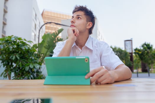 Young man in white shirt sitting at an outdoor table with a tablet and a cup of coffee, looking thoughtful and focused in a modern city environment, symbolizing concentration and digital lifestyle.