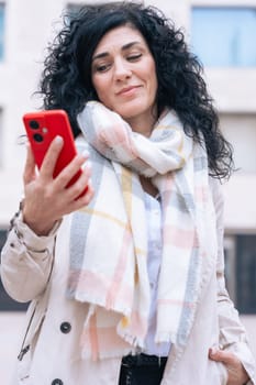 Serious caucasian woman standing outdoors in urban setting, wearing coat and scarf, using her red smartphone. She is serious and relaxed, enjoying a casual moment on a cold day in the city. Vertical