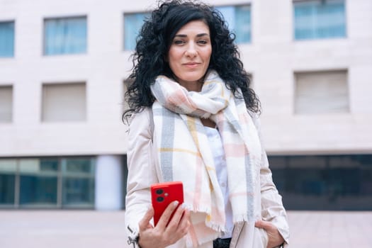 Happy caucasian woman standing outdoors in urban setting,wearing coat and scarf,using her red smartphone.She is smiling and relaxed,enjoying a casual moment on a cold day in the city looking at camera