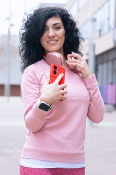 Individual portrait of a Caucasian woman in a pink hoodie smiling at the camera. She holds a red smartphone and wears headphones around her neck in a casual urban outdoor setting.Vertical